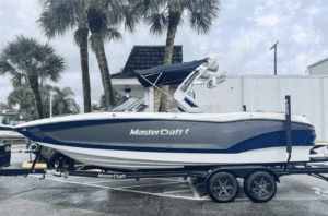 A blue and white 2023 MasterCraft X24 boat parked in a parking lot, available for sale.
