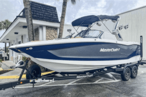 A blue and white boat, the 2023 MasterCraft X24, parked in a parking lot.